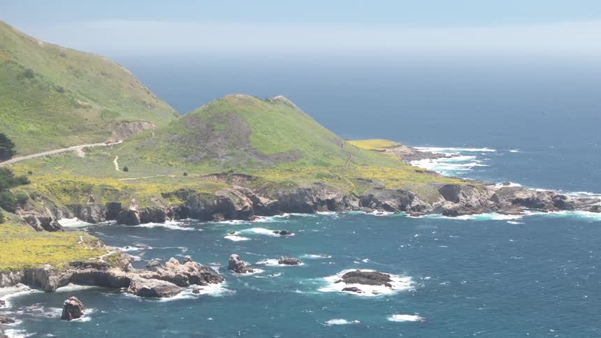 An aerial view of the pacific coastline shows the water crashing against the rocks the landscape is hilly and covered in grass and small bushes