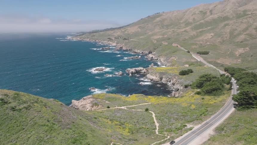 Vehicles travel along winding road between green hills and rocky cliffs facing the blue ocean