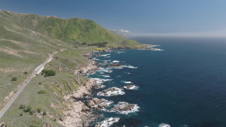 A coastal road by the rocky shoreline with ocean waves hitting the shore and a mountain backdrop at midday