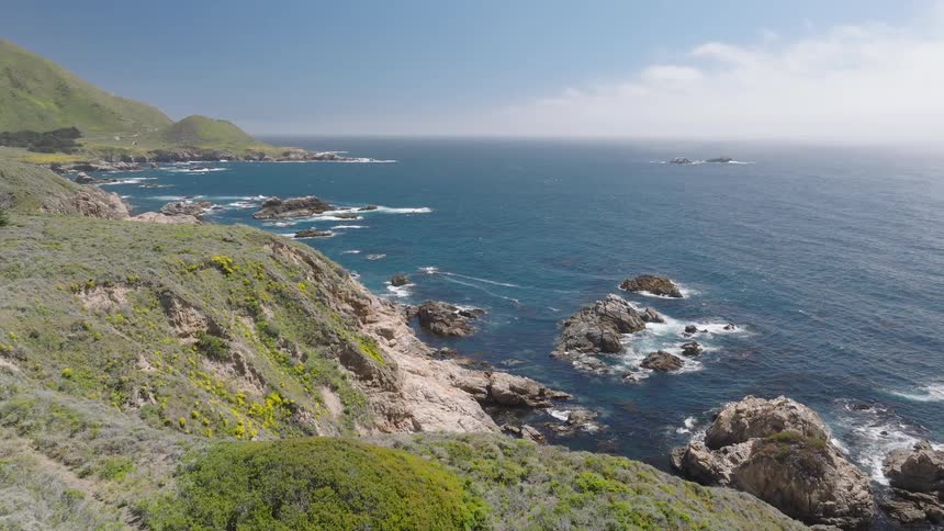 Waves crash on the rocks on the shoreline and the ocean is next to a large hill covered in grass