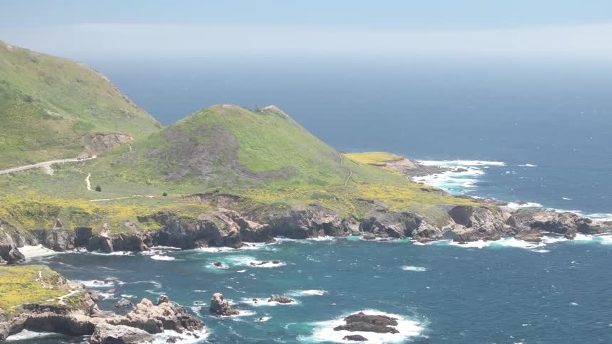 Coastal road curves along flowered and rocky slopes facing ocean stirred by white waves