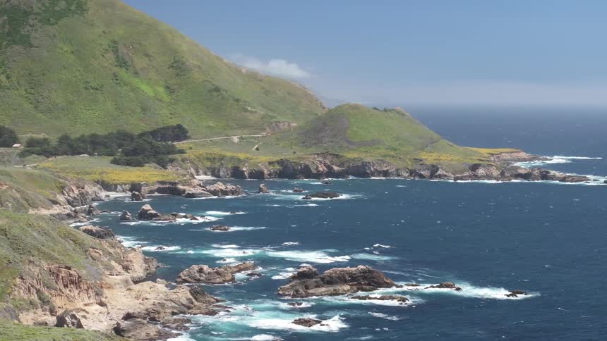 Waves crash on the rocks on the shoreline and the ocean is next to a large hill covered in grass in a sunny day