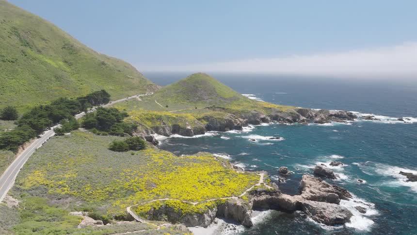 Winding road runs along hillside covered with vegetation and flowers bordering rocky coastline washed by blue ocean