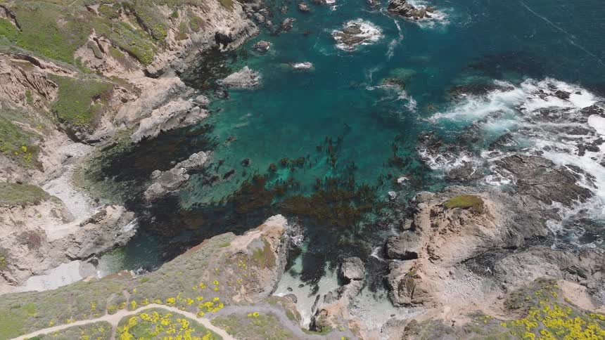 The camera moves over a cliff and shows a rocky inlet from a high angle