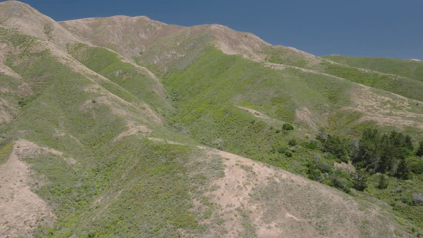 A rough mountain terrain covered in green vegetation at midday with sunlight highlighting the uneven slopes and dense foliage across the rugged landscape