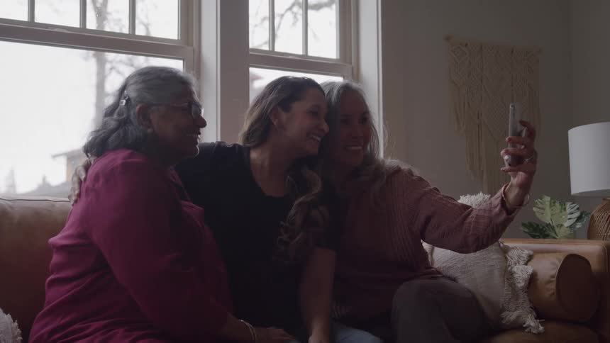 Three women sit on a couch and pose for a selfie in a living room setting
