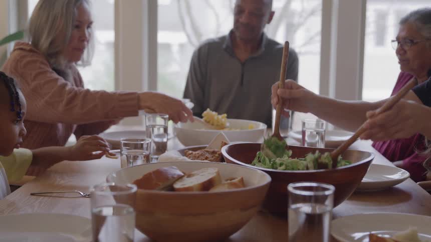 People share a meal at wooden table with assorted dishes in room lit by large windows
