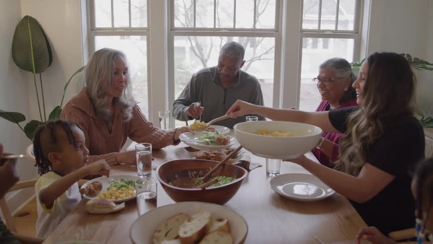 People share a meal at wooden table in front of large windows with natural light