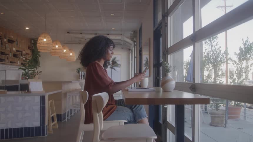 Woman sitting alone in modern caf uses phone with open laptop on high table