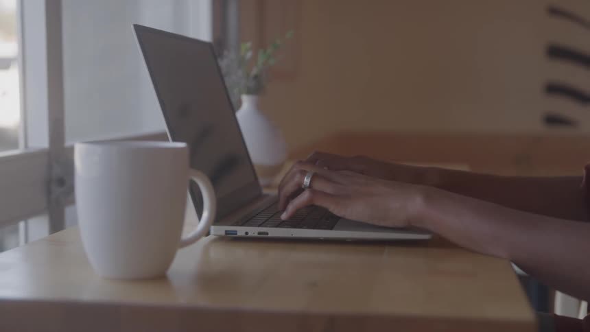A woman sitting behind a desk near a window at midday typing on a laptop with a mug placed beside her sunlight streams through the window softly illuminating the workspace