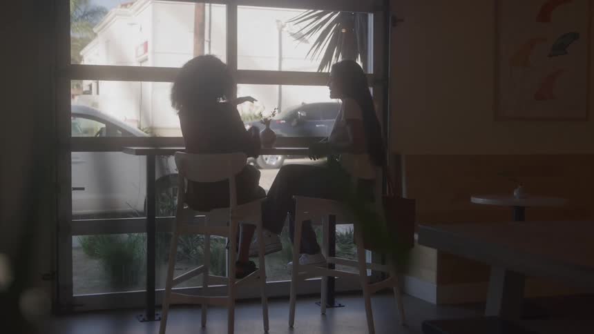 Two women are silhouetted against a large glass window as they sit and talk at a hightop table in a dimly lit cafe