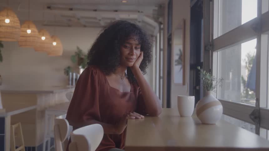 A young black woman with long black curly hair wearing a brown dress sits in a diner at midday a cup of coffee rests on the table in front of her as sunlight filters through the windows