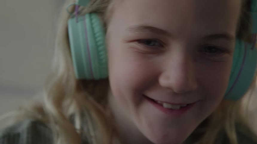 A close view of a girl with blonde hair wearing turquoise headphones smiles happily while looking down at a screen