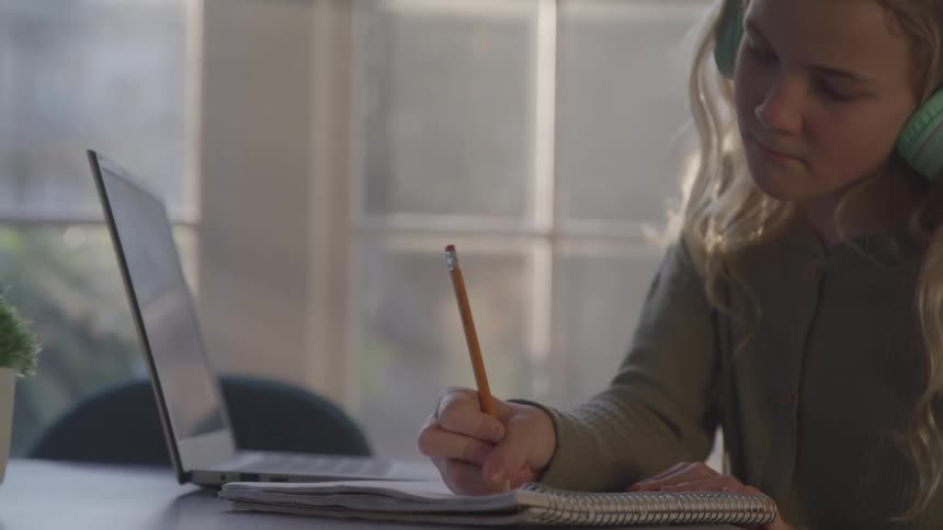 A teenage girl wearing a headset sits at a dining table at midday writing with a pencil in a book while a laptop rests open in front of her sunlight filters through nearby windows casting a warm glow over her focused workspace
