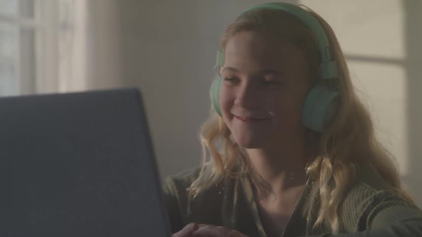 A young woman with blonde hair wearing green headphones smiles and waves at the screen while participating in a video call on a laptop in a sunlit room
