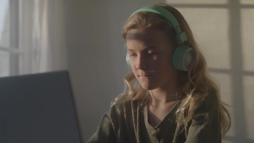 A young woman with long blonde hair wearing green headphones is focused intently on a laptop screen with natural window light illuminating her face