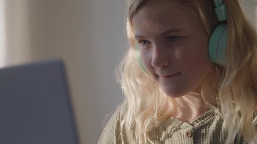 A young blonde girl wearing mint green headphones smiles while looking at a laptop the shot is a mediumcloseup with the light from the screen illuminating her face