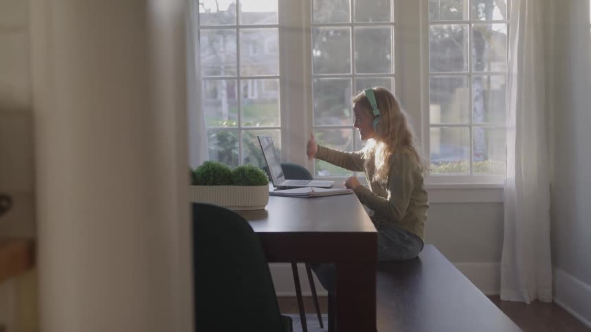A girl sits at a table and waves at her computer screen