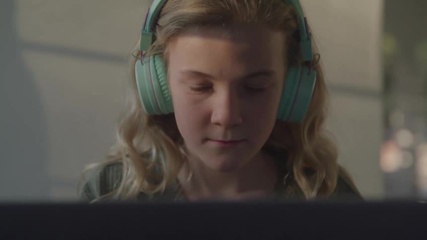 A blonde girl wearing mint green headphones intently watches something on a laptop she looks focused with the glow from the screen reflecting on her face