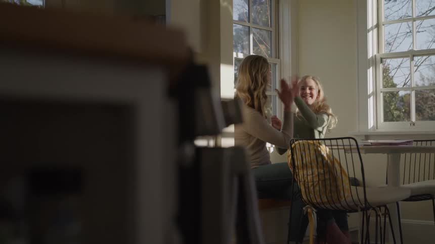 A young woman highfives her child at the dining table while working on an assignment together at midday both look excited and energized with sunlight streaming through nearby windows