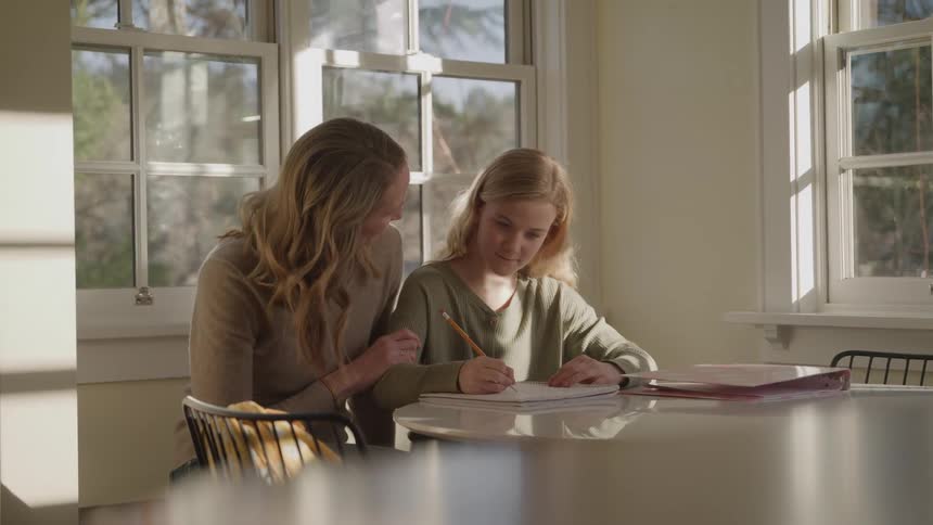 A person and a child sit at a table and the woman is teaching the young girl how to do her homework