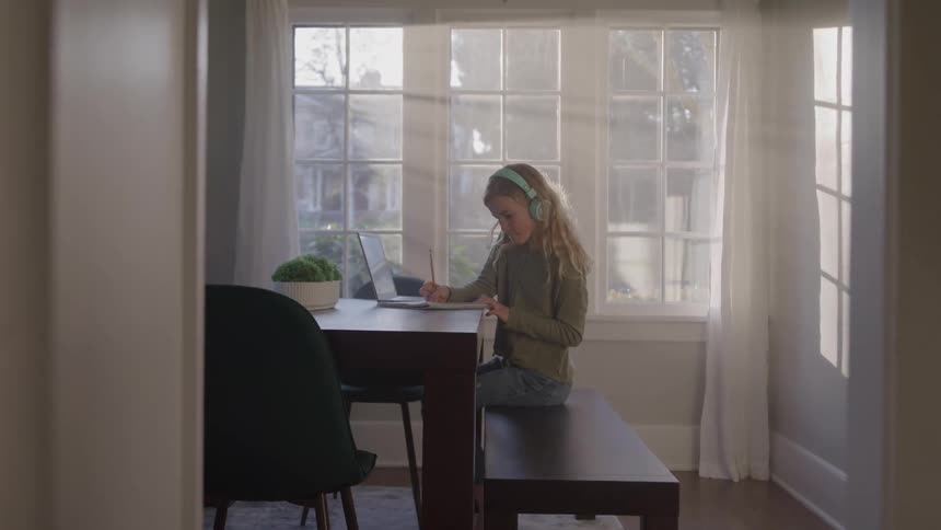 Child wearing headphones studies in front of a laptop in a sunlit room