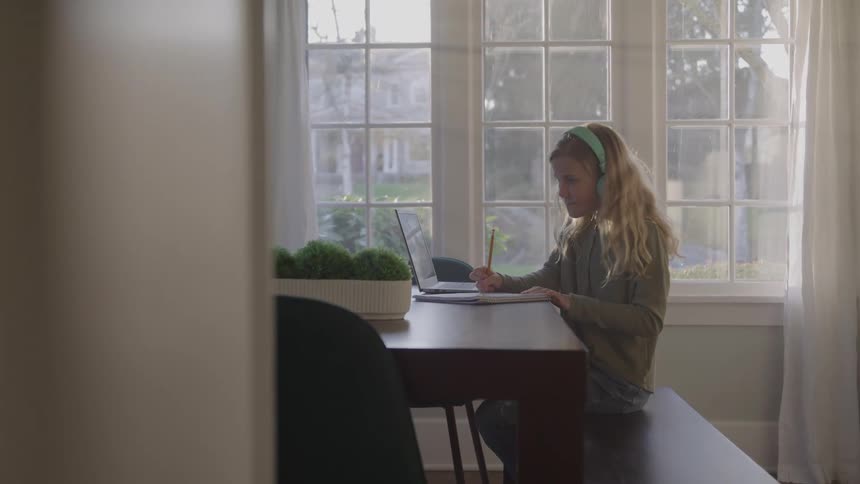 A child sits at a table near a large window writes in a notebook and works on a laptop