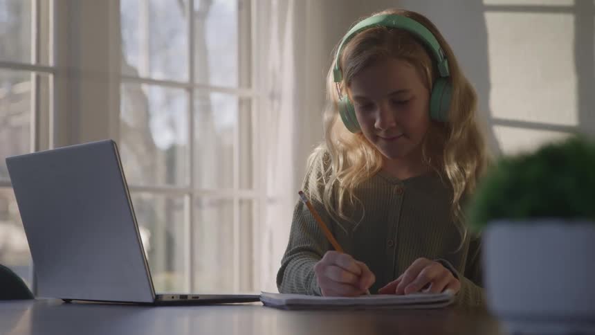 A teenage girl sits at a table near a window wears headphones and writes in a notebook with a pencil a laptop is open next to the notebook
