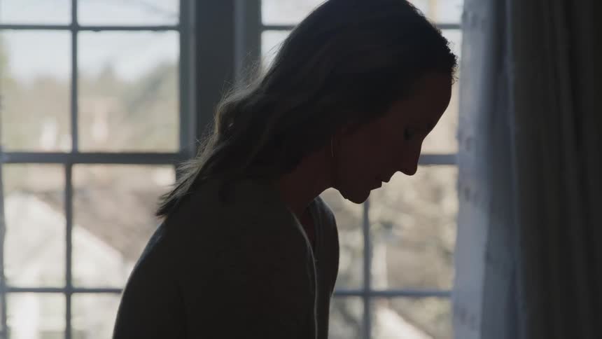 A silhouette of a woman in a dark room stands facing a bright window slowly lowering her head and looking down with a somber expression