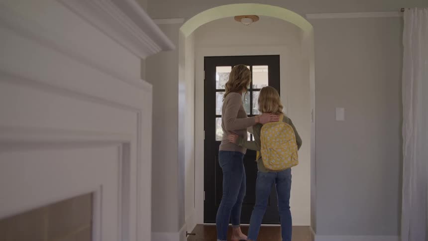 A woman wearing jeans and a sweater helps a young woman put on a yellow patterned backpack gives her a hug and watches her walk out the front door