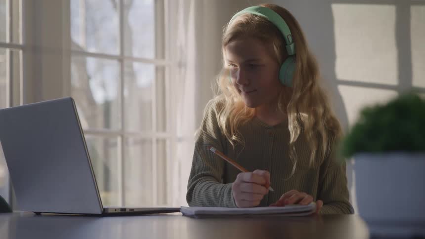 A closeup shot captures a young blonde girl wearing a green sweater and mint green headphones she is sitting at a table with a laptop open and a notebook in front of her and she looks down to write something