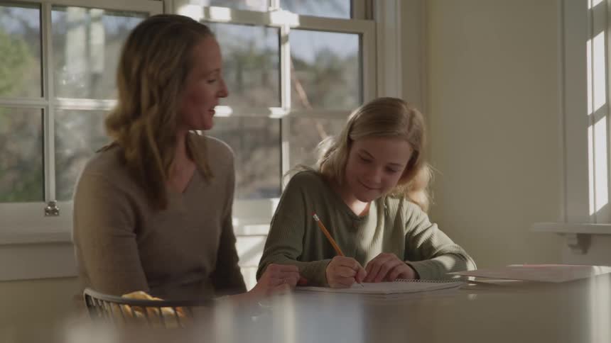 A smiling woman helps a young woman with her homework at a table in a bright room celebrating with a high five after the younger woman finishes writing with a pencil