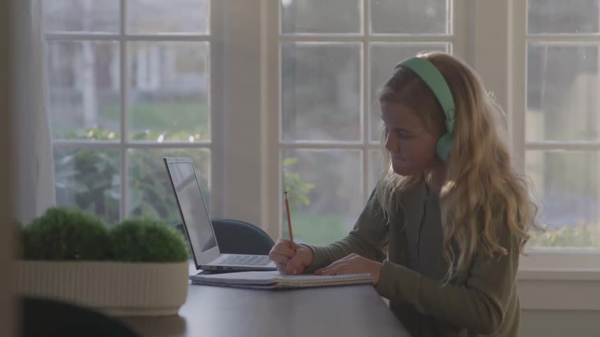 A child sits at a table near a bright window wears headphones and writes in a notebook with a pencil