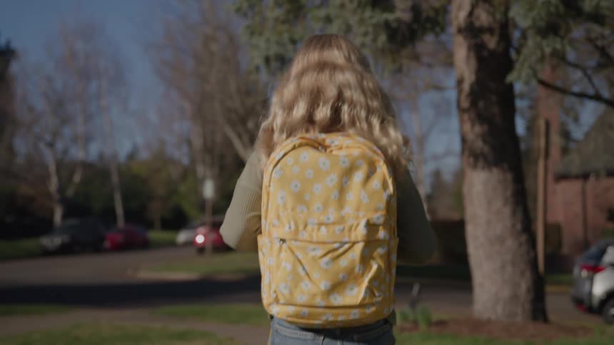 A teenage girl with long blonde hair walks on the sidewalk carrying a yellow backpack on a sunny day