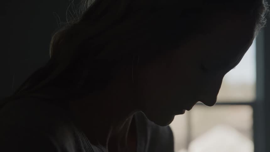 A young womans face is silhouetted in a dark room she turns her head to the side looking down with a somber or pensive expression