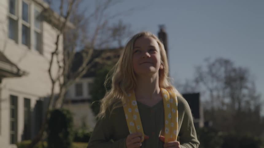 A young girl with long blonde hair and a yellow backpack stands outside a house on a sunny day she is looking up and smiling