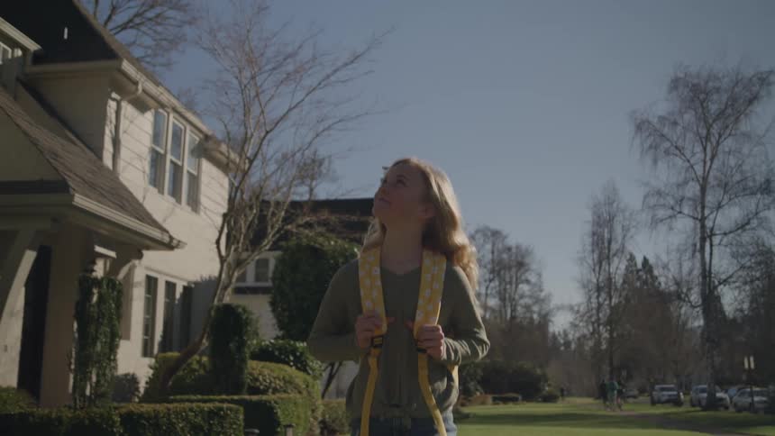 A teenage girl walks down a driveway away from a house while carrying a backpack