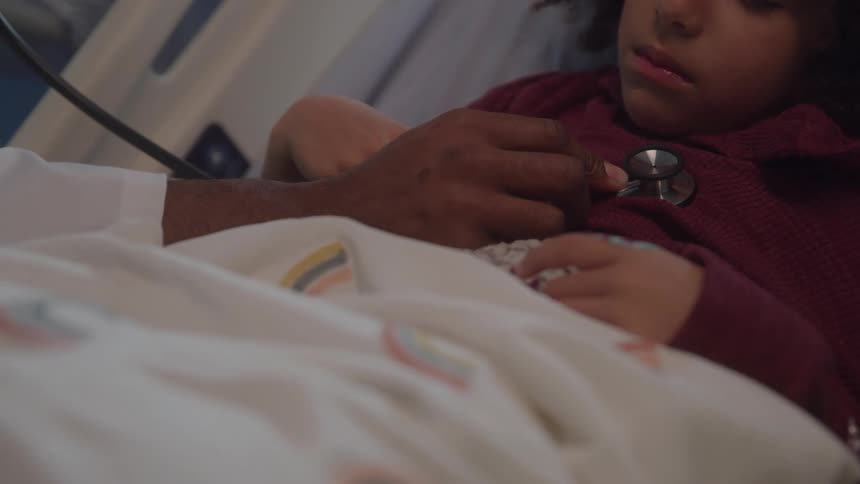 A close view shows the hand of a doctor using a stethoscope to listen to the chest of a young girl patient in a hospital bed with a rainbow patterned blanket