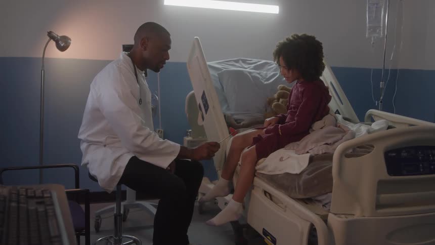 A bald male doctor in a white coat is sitting next to a young girl with curly hair in a hospital bed talking to her while gently examining her legs