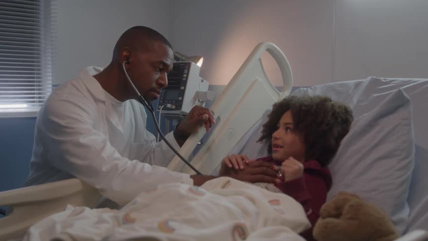 A doctor wearing a white coat uses a stethoscope to examine the chest of a young girl with curly hair who is lying in a hospital bed