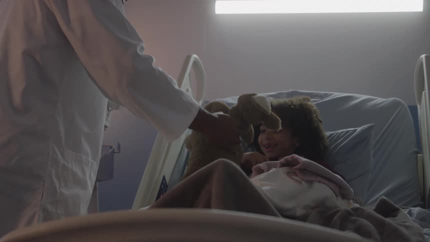 A doctor in a white coat leans over a child lying in a hospital bed holding a stuffed animal toward her