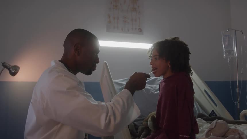 A doctor in a white coat sits beside a child on a hospital bed the doctor uses a tongue depressor to look into the childs mouth