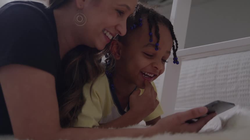 A mother and her son lying side by side on a bed at midday watching content on a mobile device both are laughing in excitement
