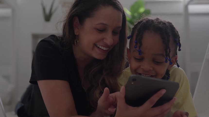 A child and mother lying together on a bed at midday both excitedly watching content on a mobile device