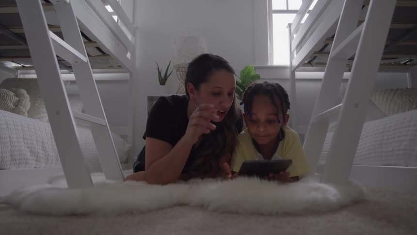 A mother and daughter look at a tablet while laying on a rug
