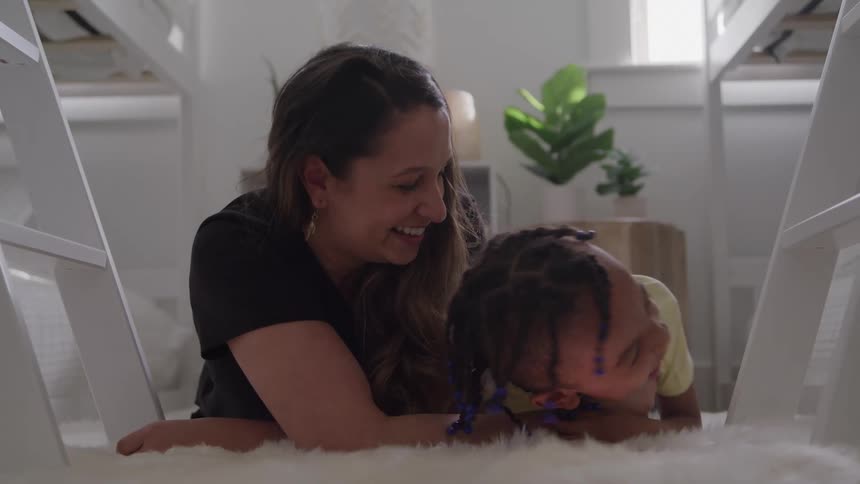 A person and a child lie on the floor between the frames of a bunk bed the two people laugh and interact closely