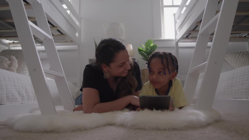 A person and a child lie on a white rug looking at and touching a smartphone