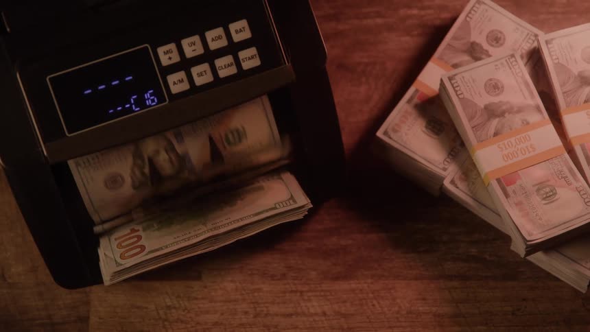 Money counting machine processing dollar bills next to stacked bundles