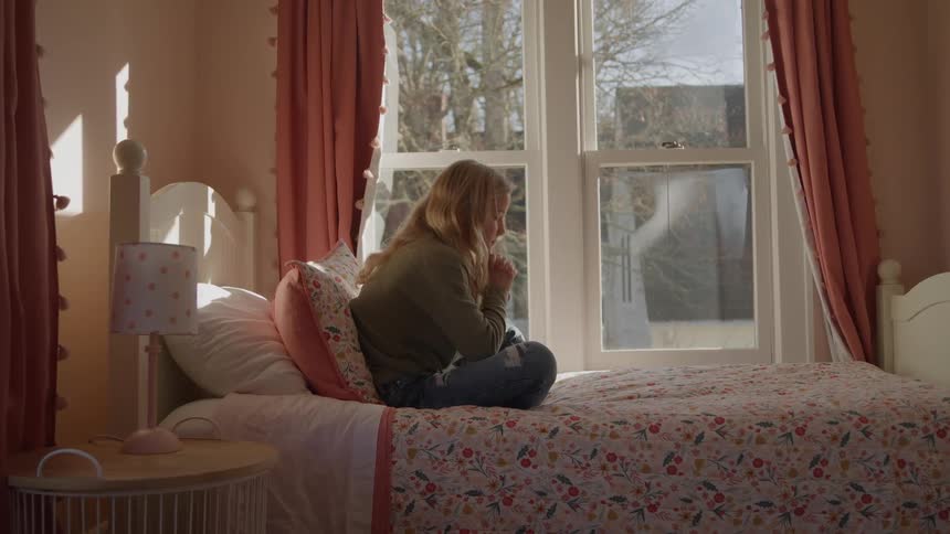 Girl sitting alone on the edge of the bed looking thoughtfully out the window