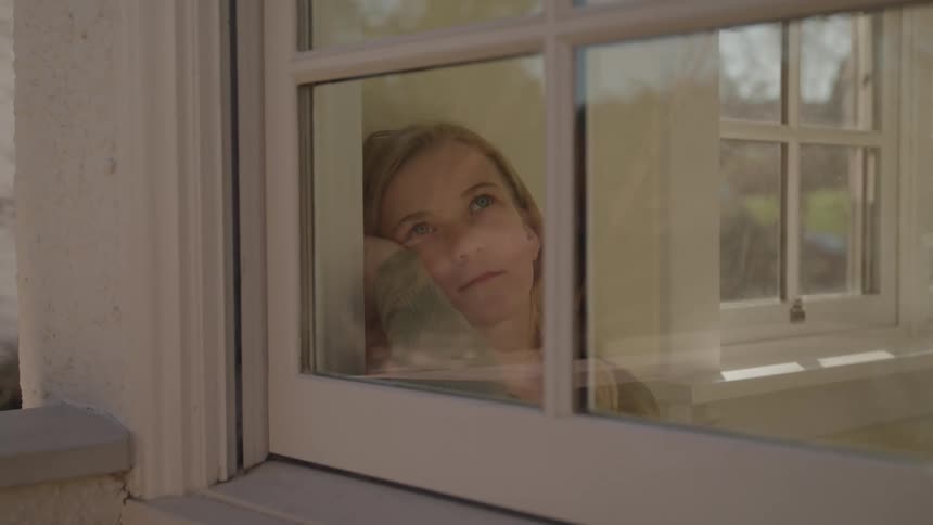 Woman looks thoughtfully out the window as sunlight illuminates her face